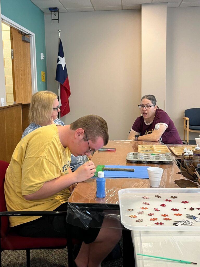 Two volunteers work with an artist at a table set with paint trays and dried leaves; one artist paints a blue and gold canvas with a foam brush.