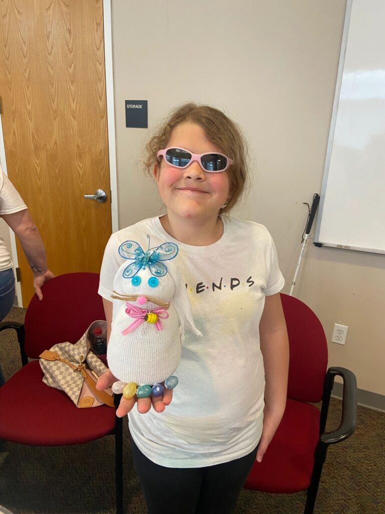 An artist holds up a finished sock bunny decorated with a blue glitter butterfly, pastel bead bracelet and pink ribbon collar, standing near a storage door.