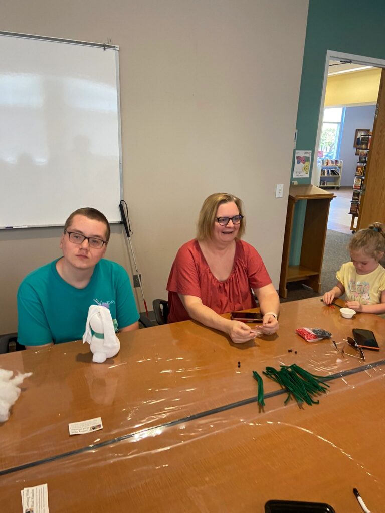 A volunteer sits with two artists at a table set with green pipe cleaners and a finished sock bunny with a ribbon bow.