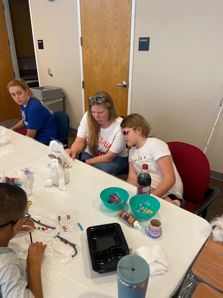 A volunteer helps an artist tie a ribbon onto a partly finished sock bunny at a table set with bowls of beads.
