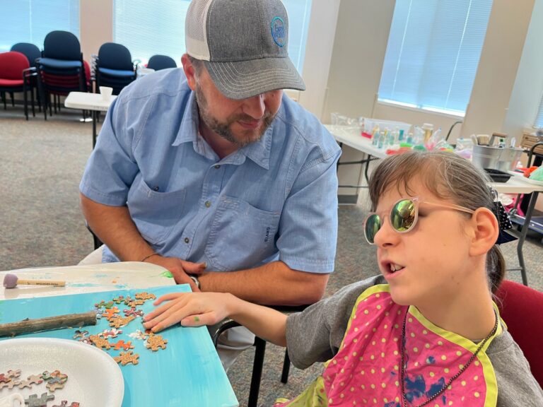 An artist glues small charms onto a blue canvas decorated with a green grass strip, a twig, and rows of dark puzzle pieces along one edge, with a yellow glitter sun.