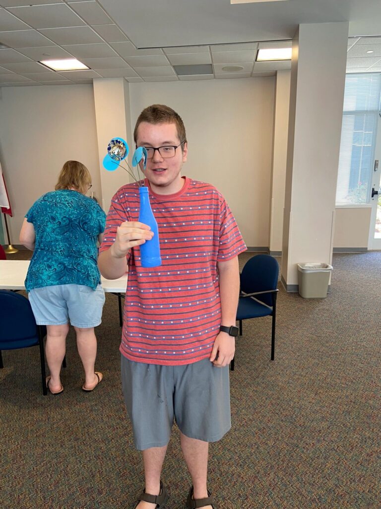 An artist holds up a blue-painted bottle with a paper flower and small charm on a wire stem, standing in the studio.