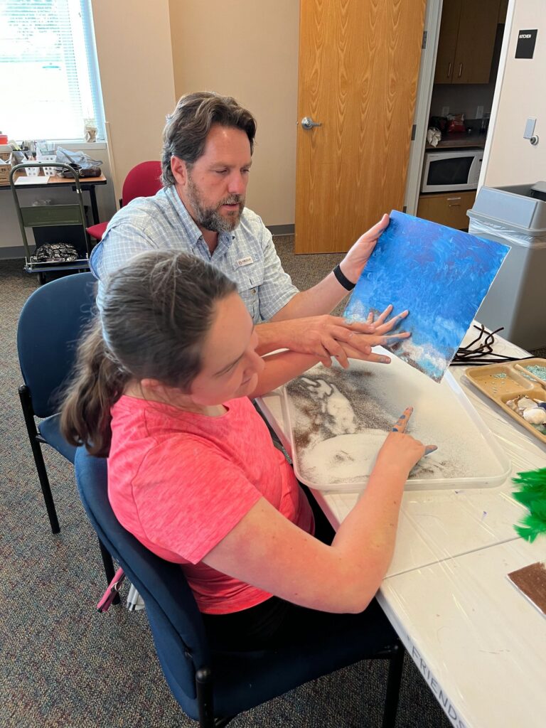 A volunteer helps an artist press their hands into sand on a blue canvas, with shells and blue gravel in a tray on the table.
