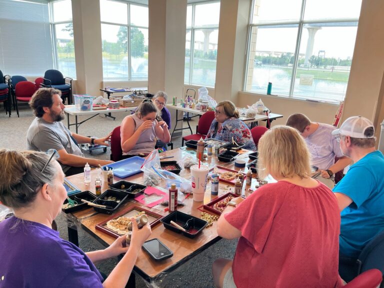 A wide view of the studio near a window overlooking a fountain, with artists and volunteers gathered around a table working on framed cork pieces.