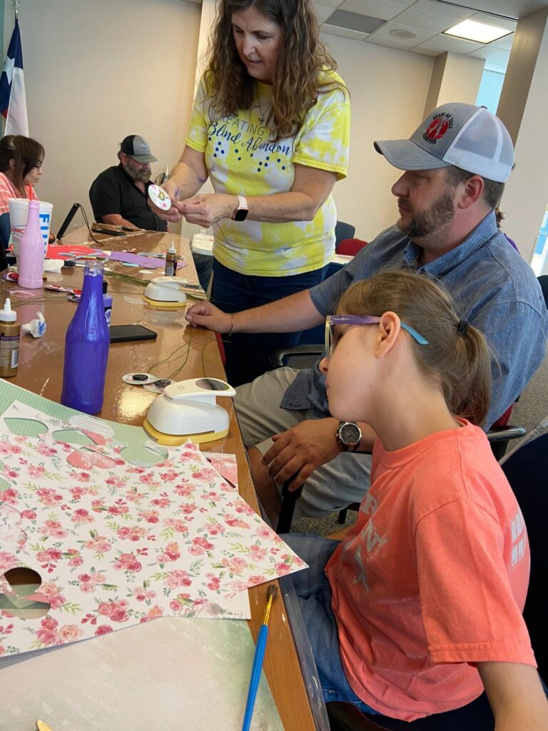 An artist and a volunteer work together at a table covered in wire stems, paper flower cutouts and paint bottles for the bottle-and-flower craft.