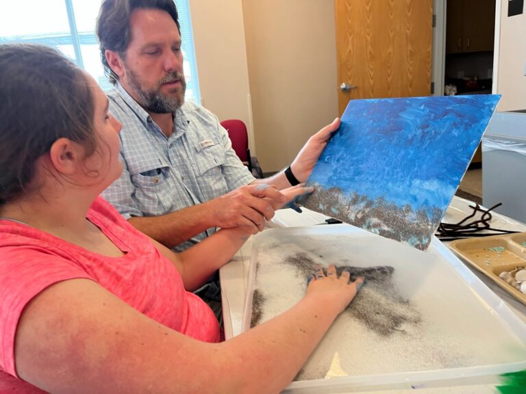 An artist and a volunteer hold up a finished blue and brown textured canvas together, with a tray of shells nearby.