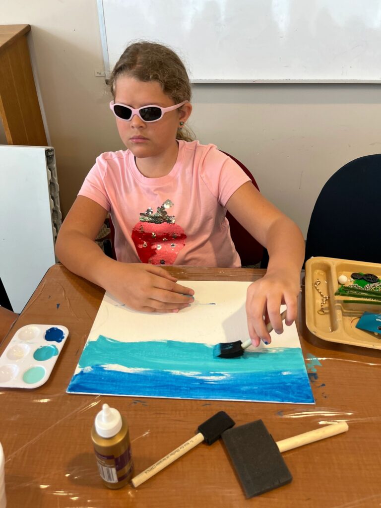 An artist paints a white canvas with a foam brush, starting a blue horizon line, with beaded strands and shells set out on a tray.