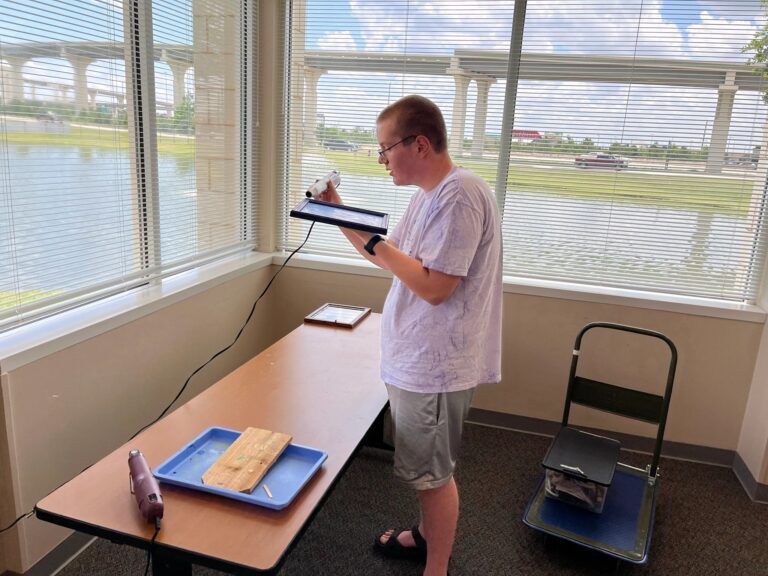 An artist uses a wood-burning tool to trace a design on a small wooden board clamped to a tray at a table by a window.