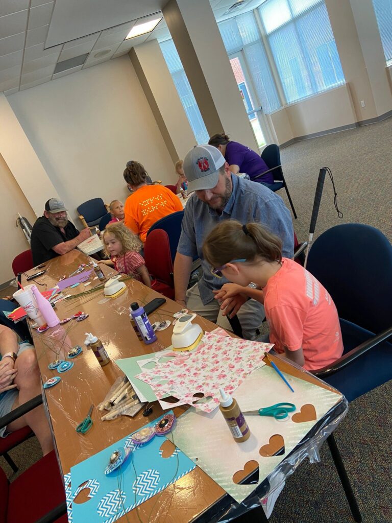A volunteer helps an artist trace a wire stem onto floral paper at a table set with scissors and paint bottles, with a white cane resting against the chair.