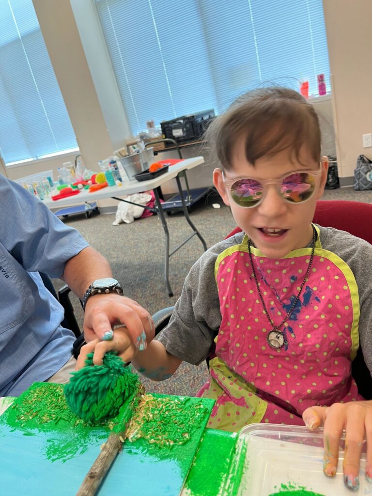 A volunteer helps an artist press a green textured pom-pom onto a canvas already dotted with painted highlights, with a paint tray beside them.