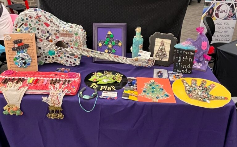 A display table set with a purple cloth, showing a range of tactile artwork: a jewelled acoustic guitar, framed mixed-media pieces, a beaded keyboard, felt gloves decorated with charms, and a sign reading Creating With Blind Abandon.