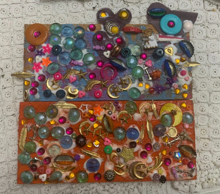 Two densely packed mixed-media boards on a lace cloth, one painted blue and pink and one orange, both crowded with glass marbles, gemstones, brass moons, stars and feathers, buttons and a small angel figurine.