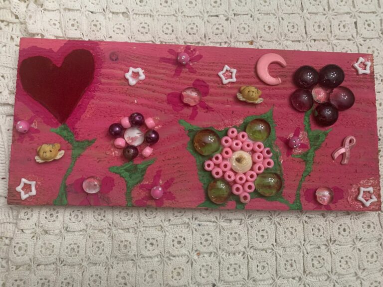 A pink-painted board decorated with a dark red heart, green painted stems, flowers made from glass gems and beads, small teddy bear charms, star charms and a crescent moon charm, with a pink awareness ribbon charm in one corner.
