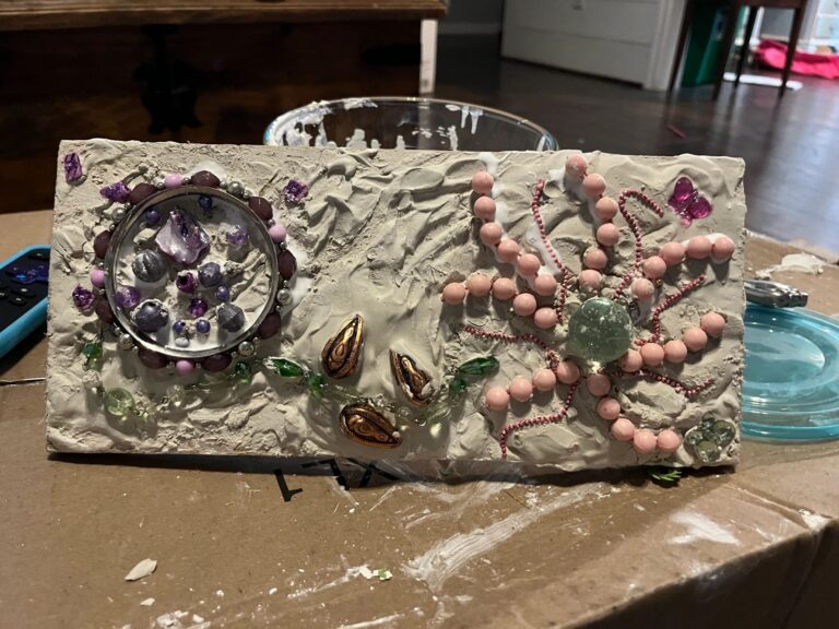 A finished Plaster of Paris board with two textured circles filled with tumbled gemstones and beads, joined by a beaded flower vine with green leaf beads and small metal charms, resting on a cardboard box.
