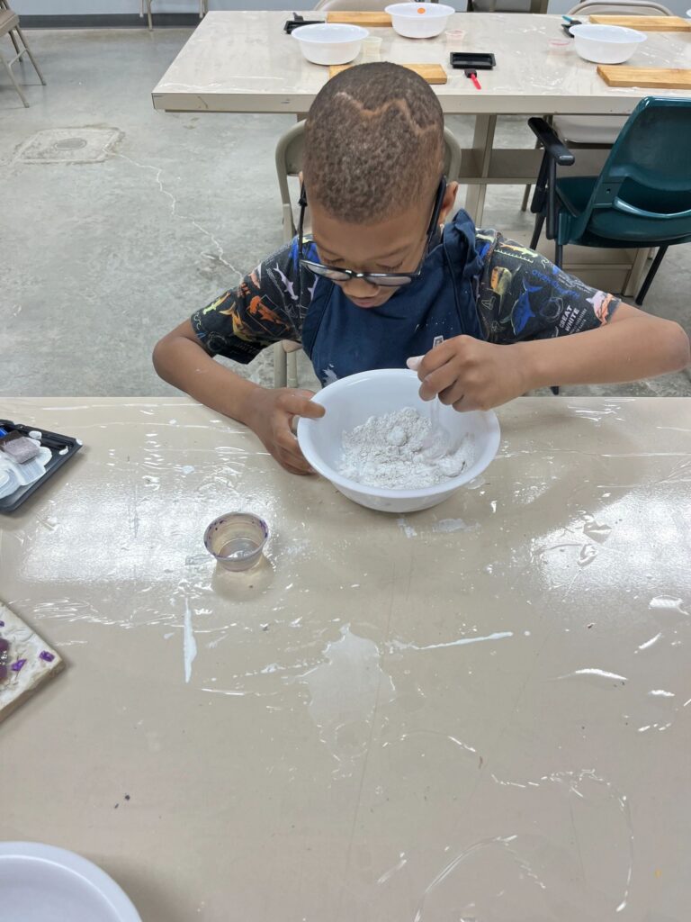An artist mixes dry Plaster of Paris powder in a bowl at the worktable, with a small measuring cup and craft supplies nearby.