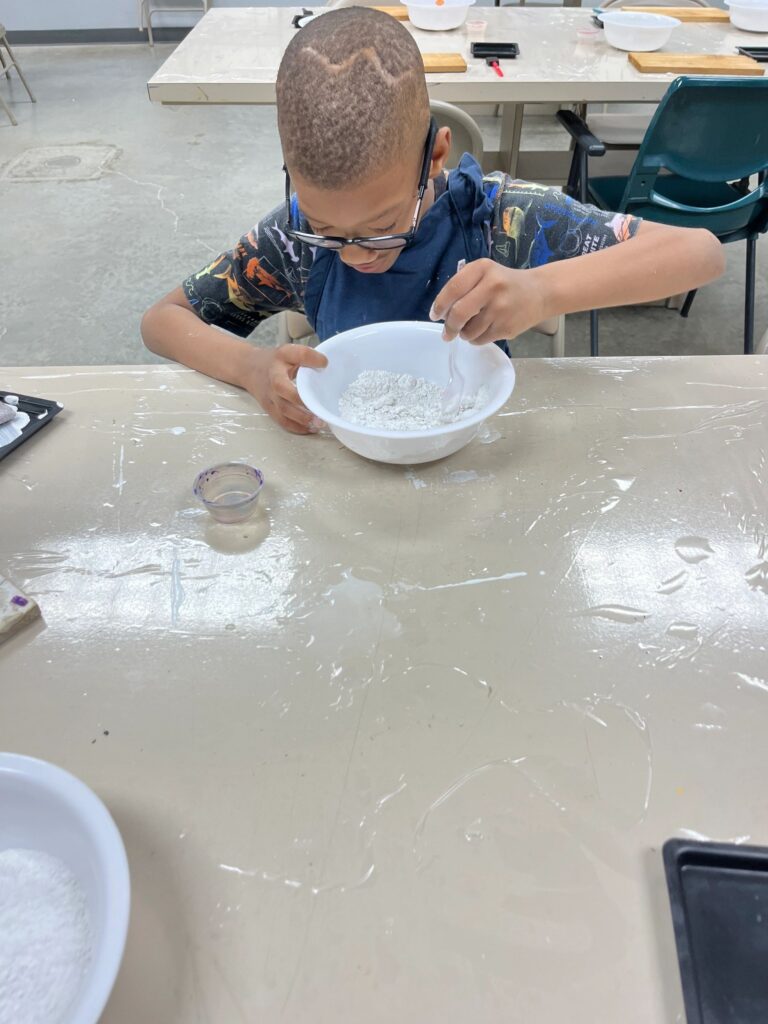 An artist stirs Plaster of Paris powder in a bowl, working carefully at the table with a measuring cup set beside them.