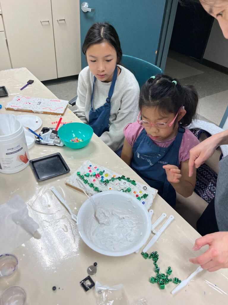 A volunteer helps two artists decorate a plaster board with green beads and small gems, with a bowl of wet plaster and colourful decorations spread across the table.
