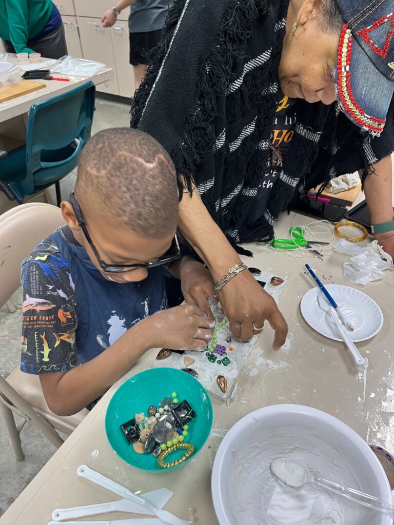 A volunteer helps an artist press beads and small charms into a plaster board, with a bowl of green beads and a bin of decorations on the table.