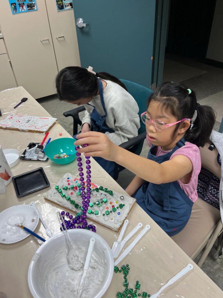 An artist pours a strand of purple beads onto a plaster board already dotted with green beads and small gems, with a bowl of wet plaster on the table.