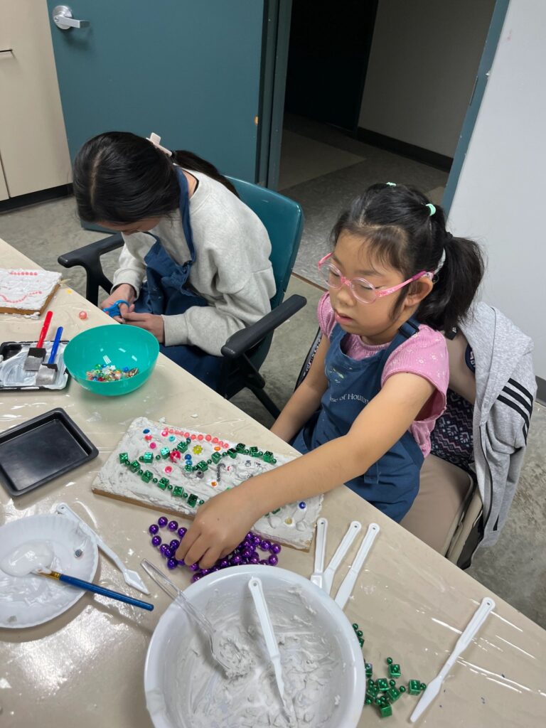 An artist arranges purple beads on a plaster board next to a bowl of wet plaster, with green beads and small charms scattered across the table.