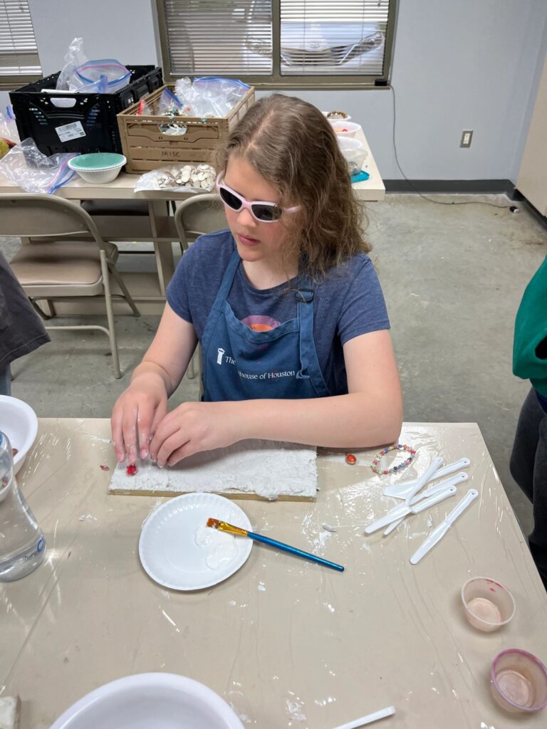 A volunteer helps an artist press beads into a plaster board, both working closely together at the table with a bowl of wet plaster nearby.