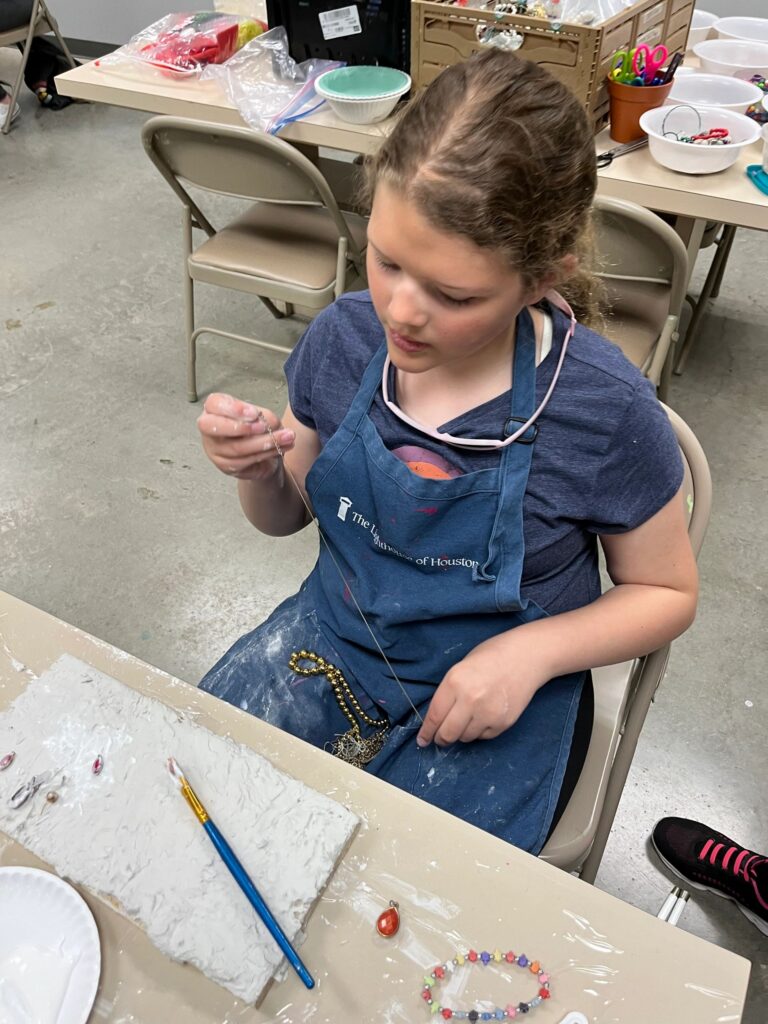 An artist works on a plaster board dotted with small gems and a red charm, with beaded bracelets and a paintbrush laid out on the table.