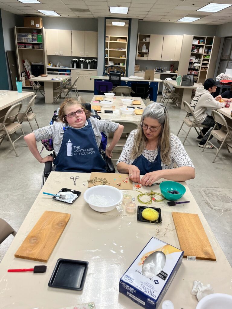 An artist threads beads onto wire, working at a table with a plaster board and a bowl of small charms and pendants nearby.