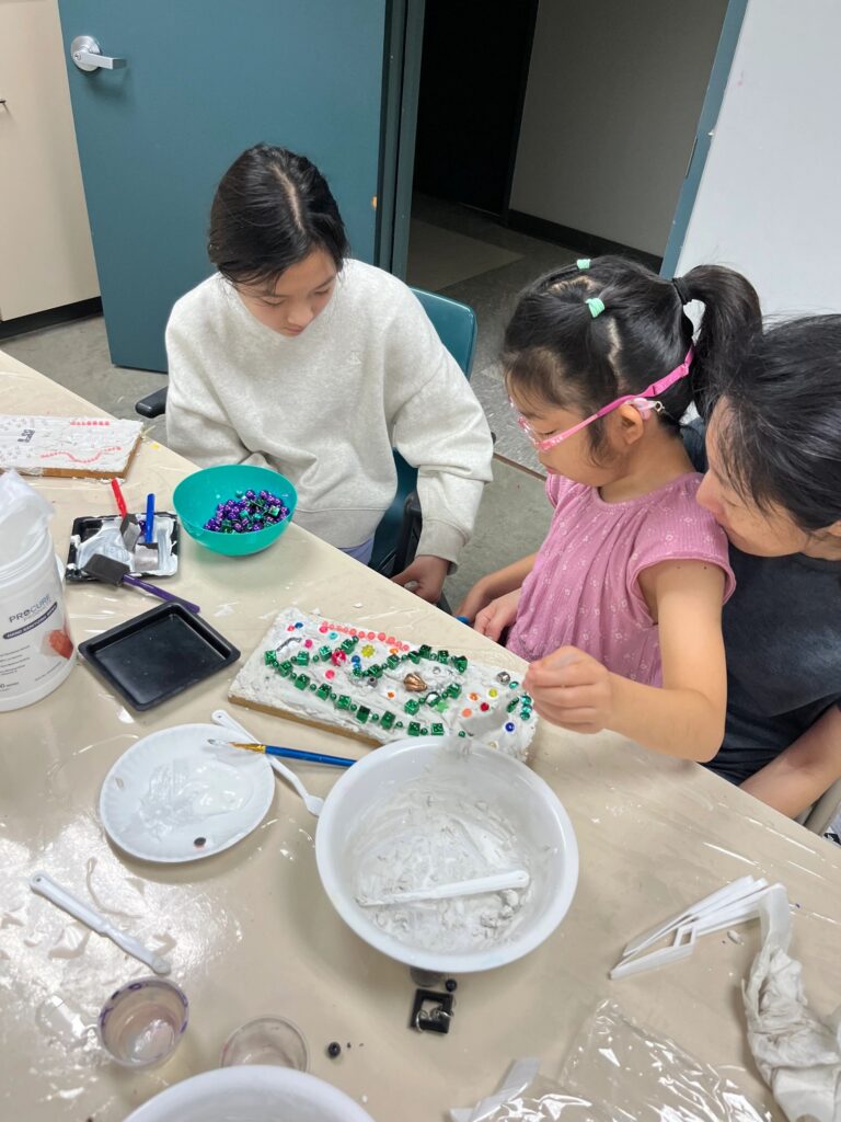 A volunteer helps two artists decorate a plaster board with green beads arranged in swirling lines, with a bowl of green beads and craft supplies on the table.