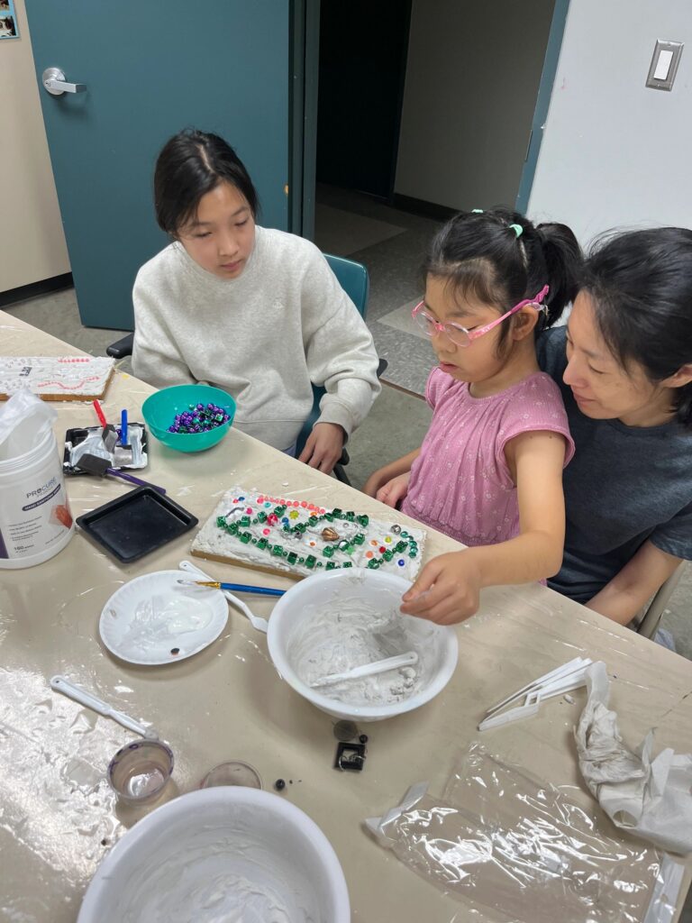 An artist presses green beads into wet plaster on a board, with a volunteer nearby and a bowl of green beads on the table.