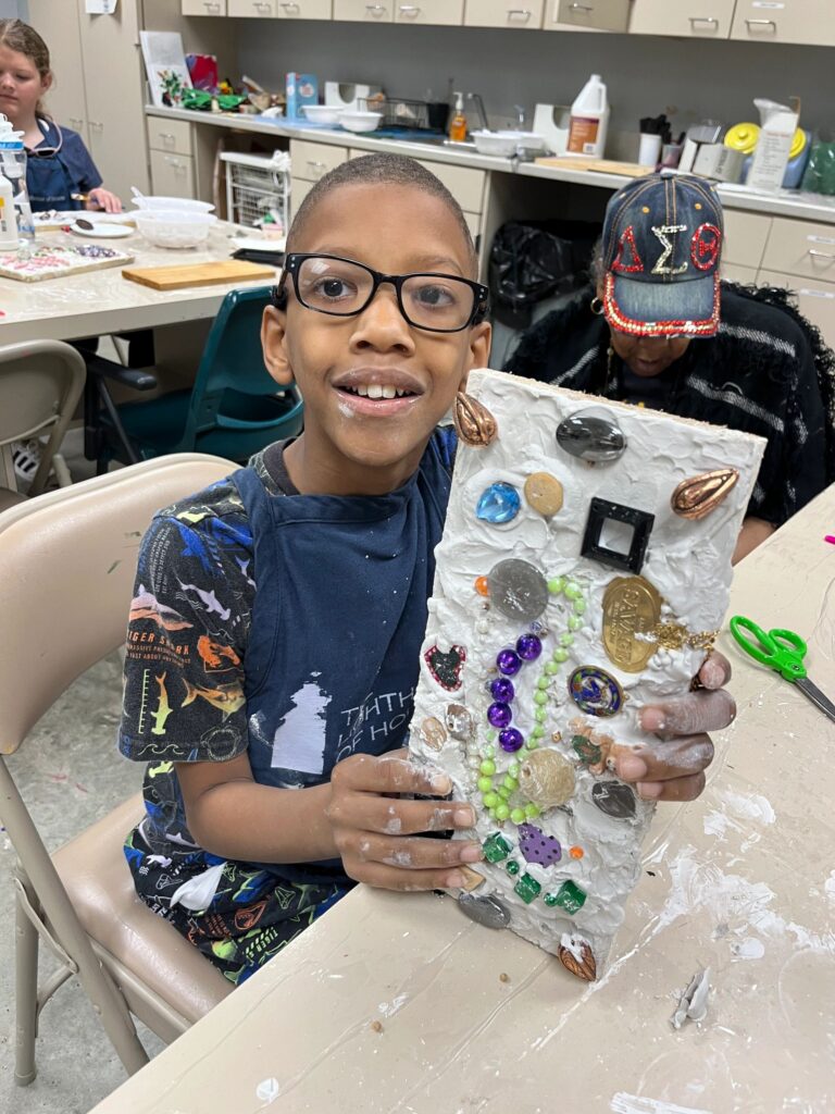 An artist holds up a finished plaster board decorated with a spiral of purple beads, gold charms and gemstones pressed into the textured surface.