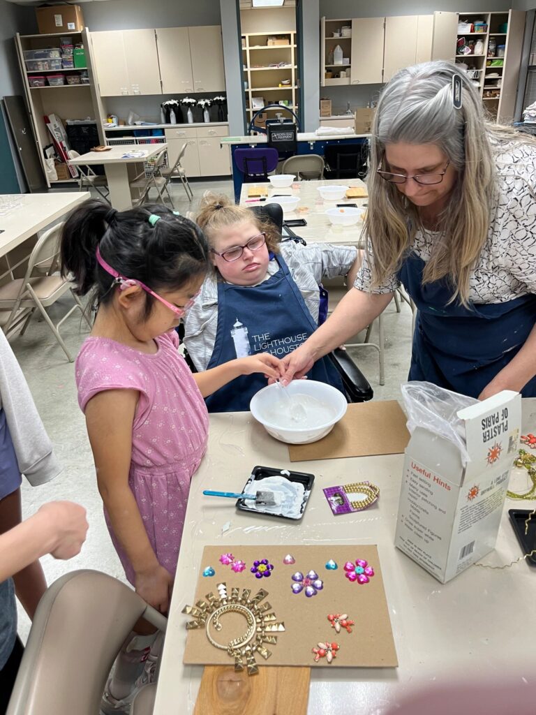 A volunteer helps an artist mix Plaster of Paris in a bowl, with a finished board decorated with gold charms and beaded flowers on the table nearby.