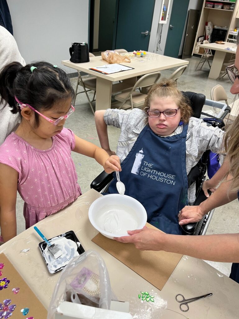 A volunteer holds a bowl steady while an artist stirs wet plaster with a spoon, seated at a table set with a plaster board and craft supplies.