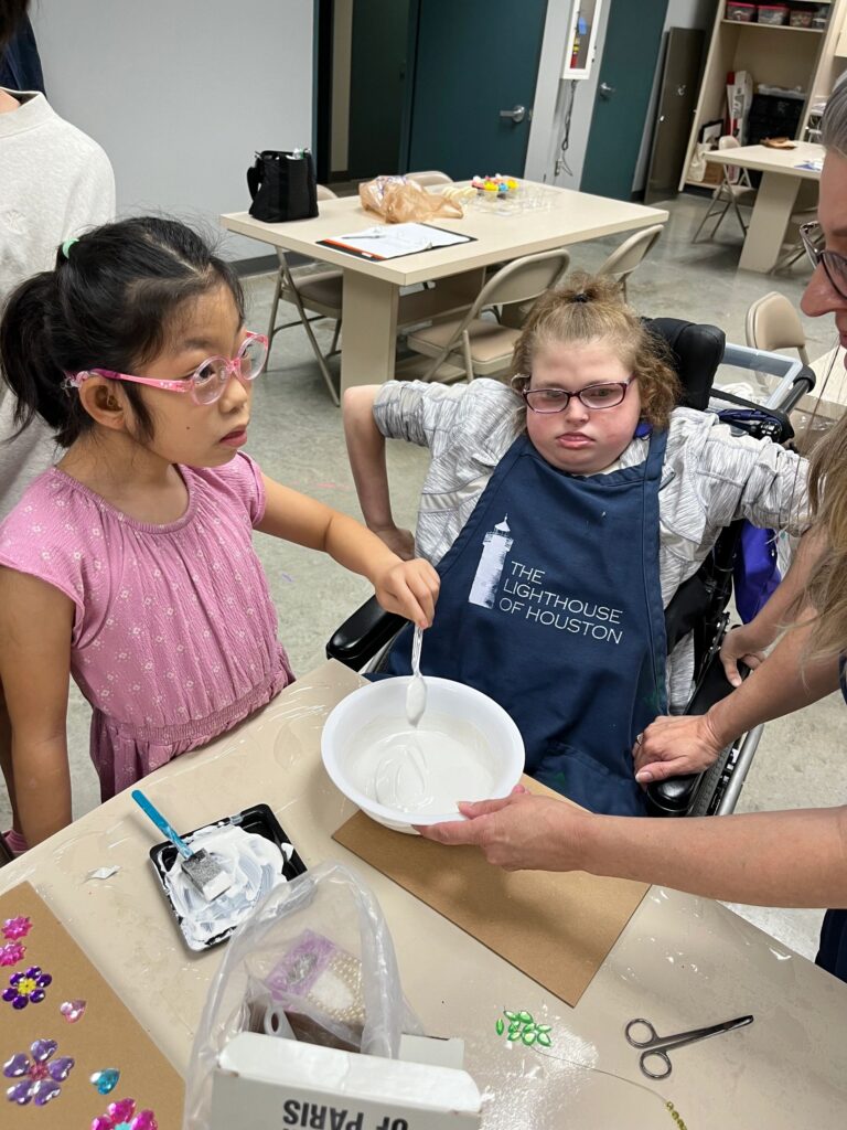 A volunteer helps an artist mix plaster in a bowl at the table, with a box labelled Plaster of Paris and beaded jewellery pieces nearby.
