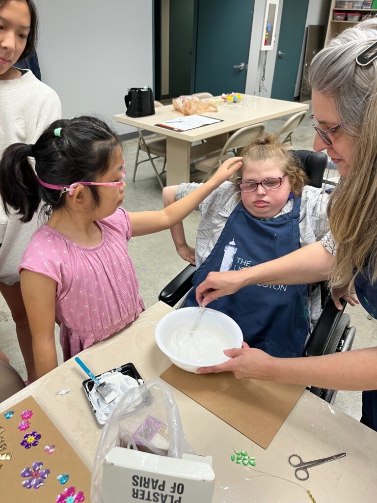 A volunteer helps an artist touch the wet plaster in a bowl, with a board and craft supplies on the table and a Plaster of Paris box in the foreground.