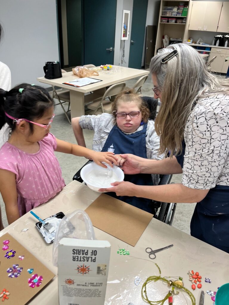 A volunteer helps an artist mix plaster in a bowl, with a board of pressed flower charms and beaded jewellery spread across the table.