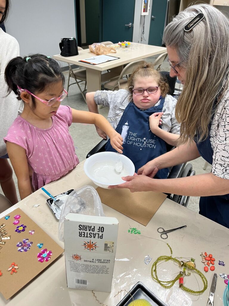 A volunteer helps an artist mix plaster in a bowl at the table, with a Plaster of Paris box and beaded jewellery pieces nearby.