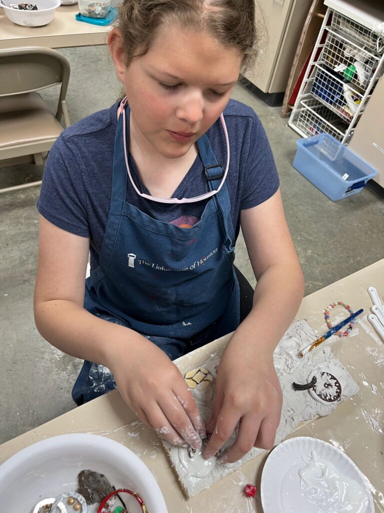 An artist presses small tiles into wet plaster on a board, with a beaded bracelet and paintbrush resting nearby on the table.