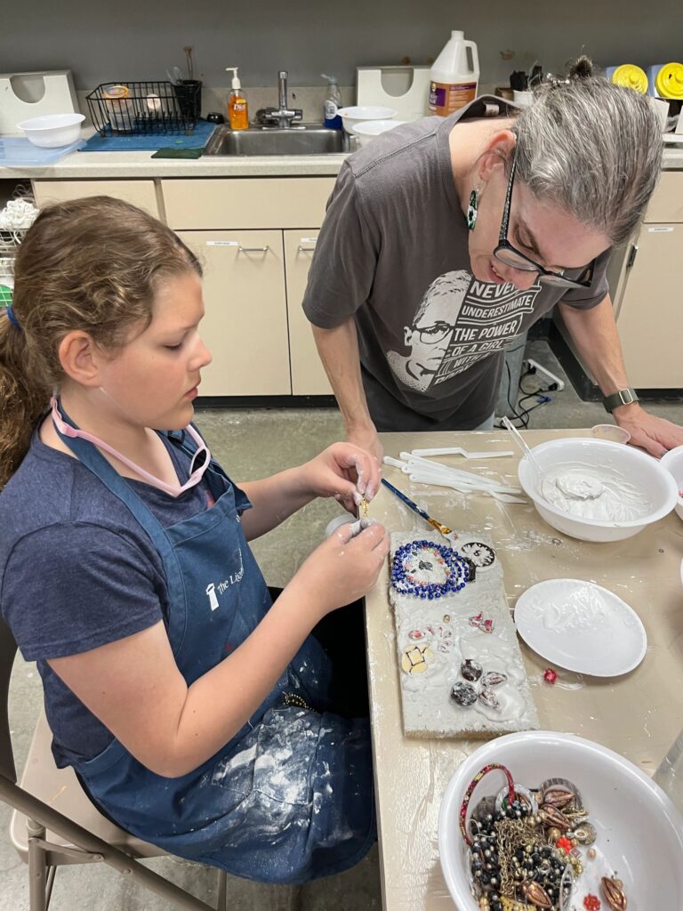 A volunteer helps an artist press blue beads and small gems into a plaster board shaped like a heart outline, with bowls of wet plaster on the table.