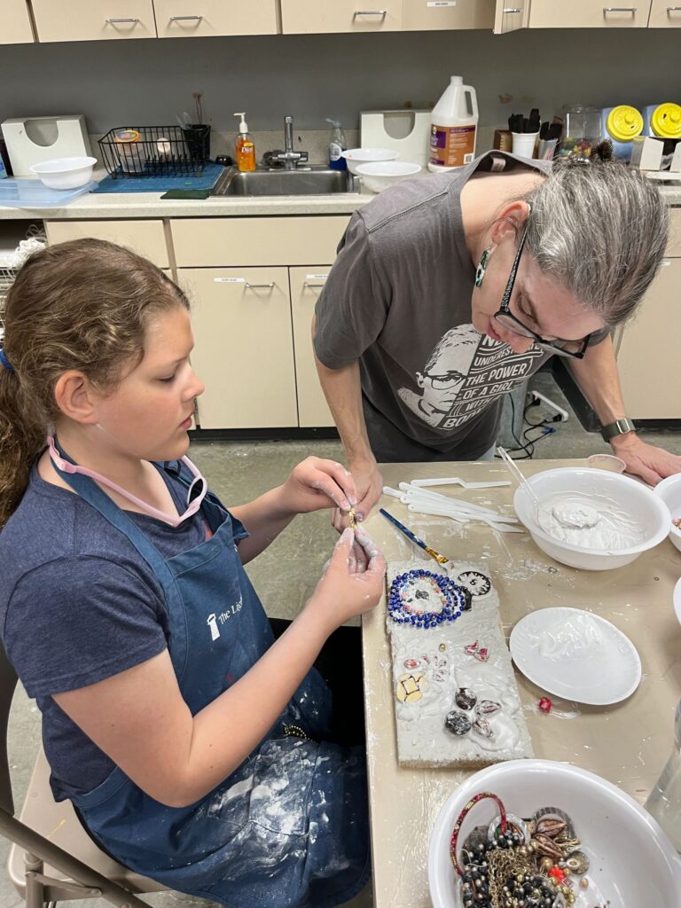 A volunteer helps an artist decorate a plaster board with blue beads arranged in a heart shape, with a bowl of wet plaster and small charms on the table.