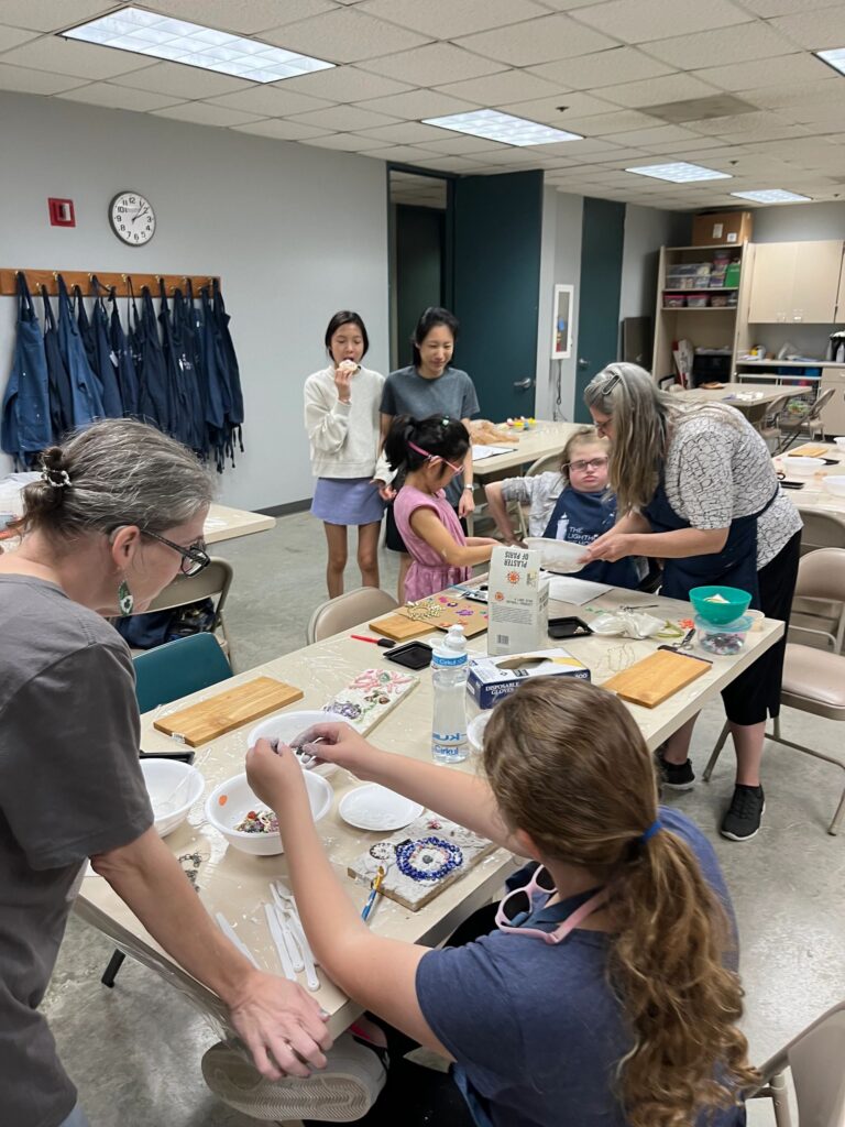 A wide view of the studio, with artists and volunteers gathered around several tables working with plaster and beads, and aprons hanging on hooks along the wall.