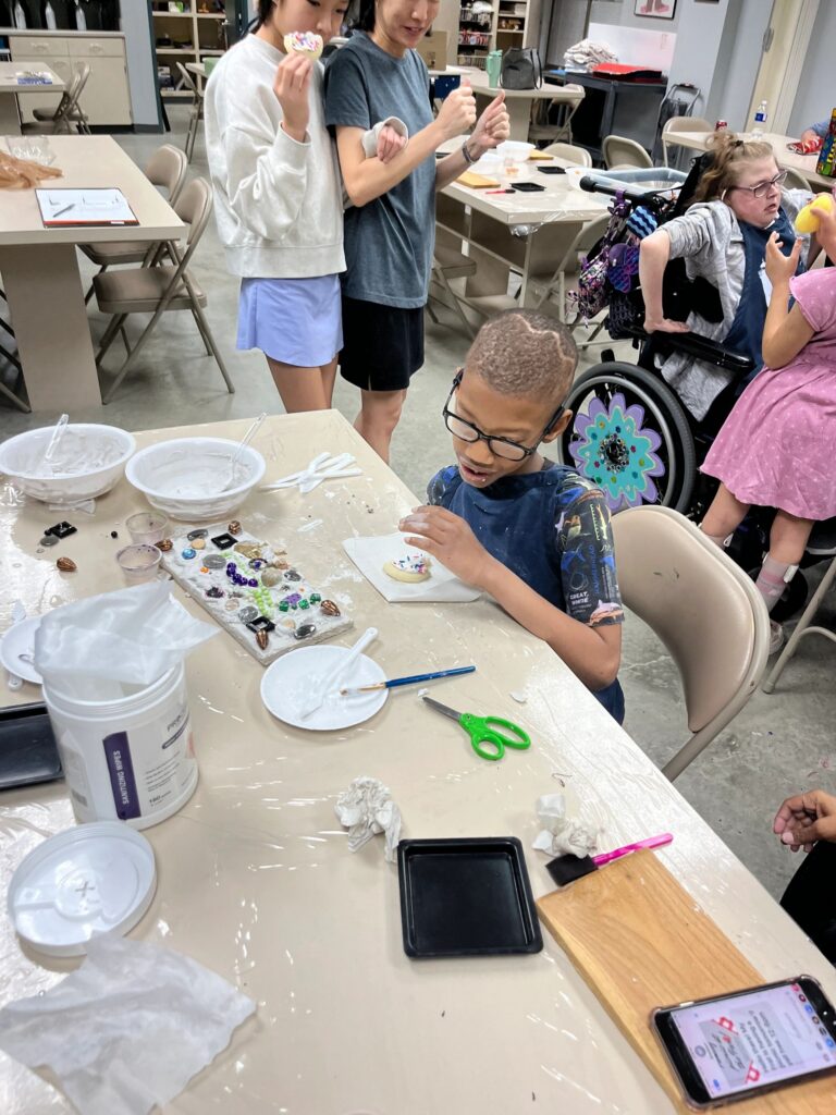 A volunteer helps an artist decorate a cookie with sprinkles at a table covered in plaster boards, bowls and craft supplies, with others eating cookies nearby.