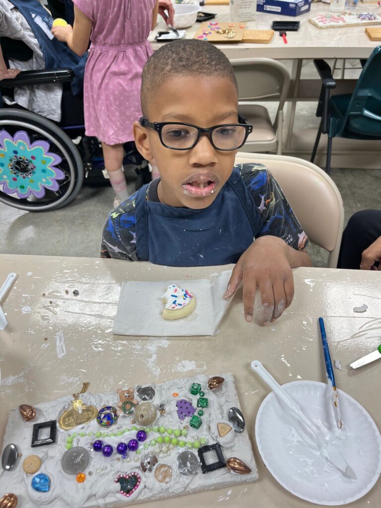 An artist sits at a table covered in puzzle-piece confetti and paint trays in red, pink and purple, with a finished heart made from pink and white puzzle pieces on a green background.