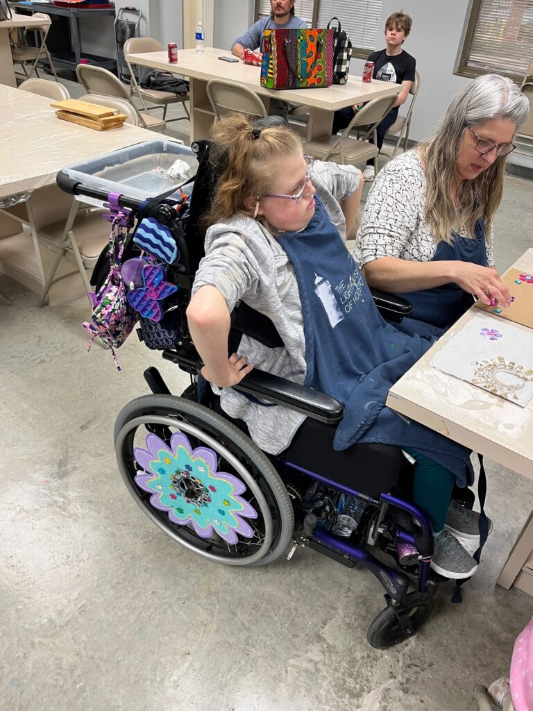 An artist paints puzzle pieces pink and purple at a table covered in newspaper, with several paint trays and loose puzzle pieces spread across the surface.