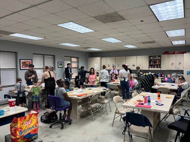 A wide view of the studio, with artists and volunteers gathered around several tables working on painted wooden boards and sorting craft supplies in bowls and trays.