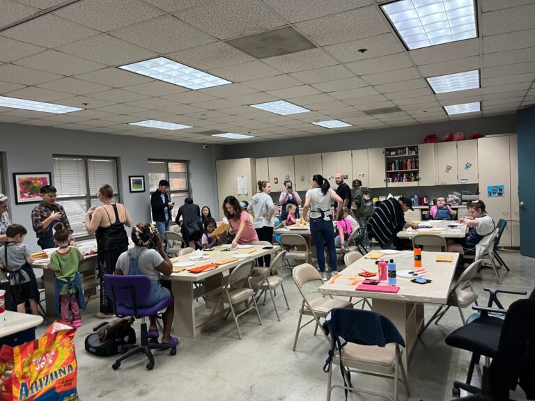 A wide view of the studio from a different angle, showing artists and volunteers seated at tables covered in paint bottles, plates and finished boards.