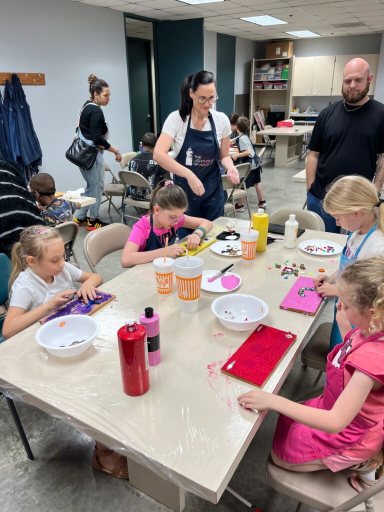 Several artists work at a shared table, pressing gems, buttons and bottle caps into freshly painted boards, with cups, paint bottles and bowls of small decorations spread across the tabletop.