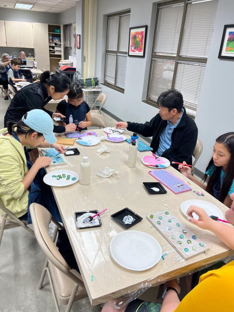 A wide view of the studio, with artists and volunteers gathered at several tables, sorting decorations into plates and working on boards painted blue, pink and yellow.