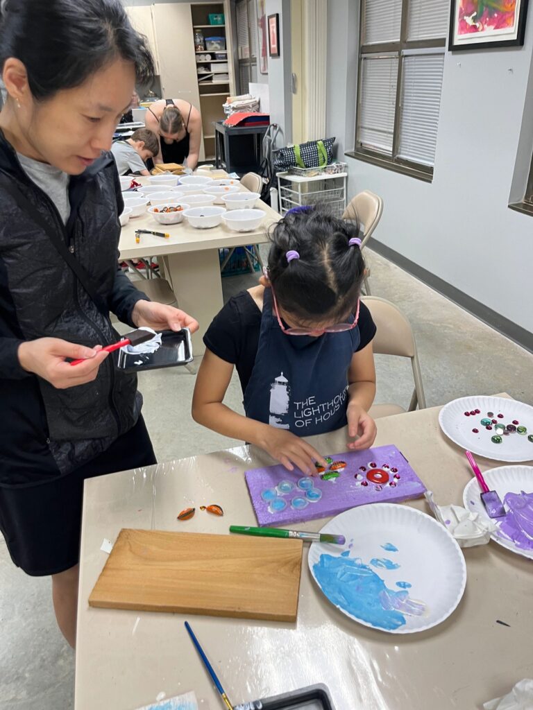 An artist presses glass gems into wet paint on a purple-painted board, with a volunteer helping steady the piece and plates of blue and pink paint nearby.