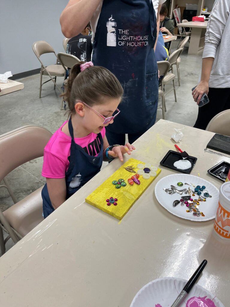 A volunteer helps an artist decorate a yellow board with bottle caps and glass gems, with a plate of extra decorations set within reach.
