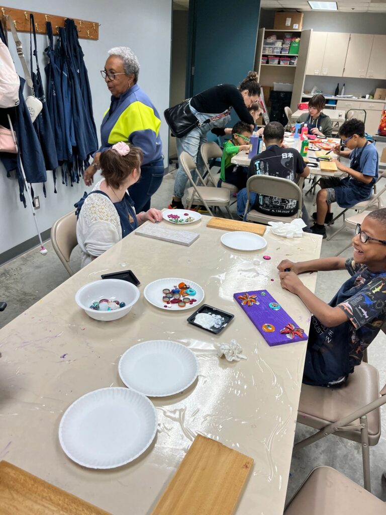 A volunteer holds a plate of decorations while an artist presses beads and gems onto a purple board, with bowls of glass gems and gold charms on the table.